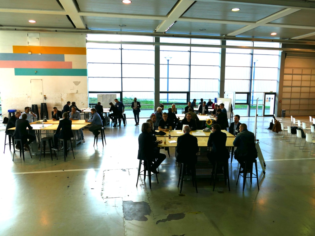 Mehrere Gruppen mit ca. 10 Personen sitzen um Tische in einer großen Halle und diskutieren